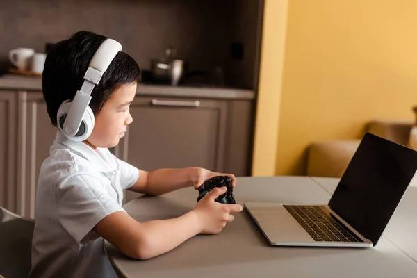 KYIV, UKRAINE - APRIL 22, 2020: Chico asiático jugando video juego con joystick y laptop con pantalla en blanco en cuarentena. - foto de stock