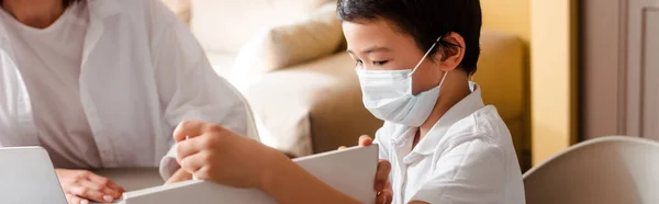 Asian boy in medical mask studying with mother at home on quarantine, horizontal concept — Stock Photo