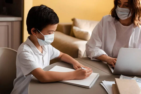 Cute asian boy in medical mask studying with mother at home on quarantine — Stock Photo