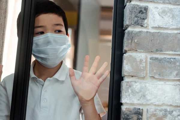 Sad asian boy in medical mask looking through window on quarantine — Stock Photo
