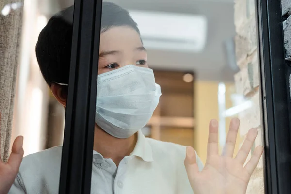 Asian boy in medical mask looking through window on quarantine — Stock Photo