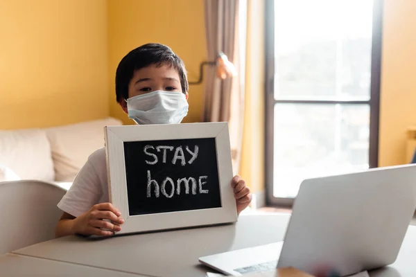 Азіатський хлопчик в медичній масці тримає дошку з перебуванням вдома, вимотуючи перед ноутбуком під час самоізоляції — стокове фото