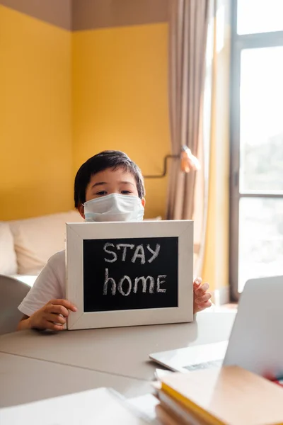 Chico asiático en la mascara médica en el portón de apoyo con carta de estancia en casa delante del portátil durante la epidemia de coronavirus. - foto de stock