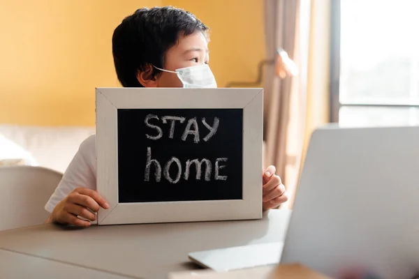 Chico asiático en mascara médica en la placa base con la estancia en casa de cartas delante del ordenador portátil durante la cuarentena. - foto de stock
