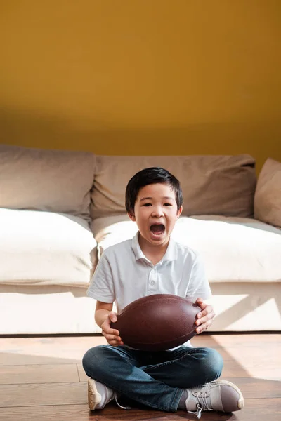 Un garçon asiatique excité avec une balle de rugby criant et regardant un match sportif à la maison en quarantaine — Photo de stock