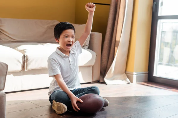 Jovencito asiático feliz con pelota de rugby gritando y viendo partidos deportivos en casa en cuarentena. - foto de stock