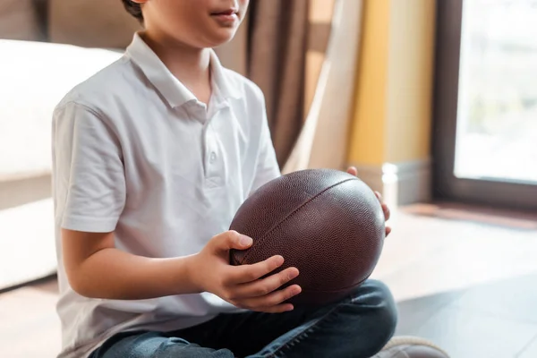 Cropped view of asian boy with rugby ball sitting on floor at home on quarantine — Stock Photo