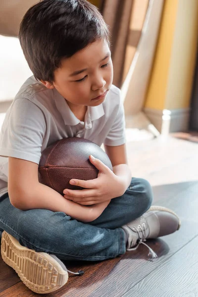 Triste ásia menino com rugby bola sentado no chão no casa no auto isolamento — Fotografia de Stock