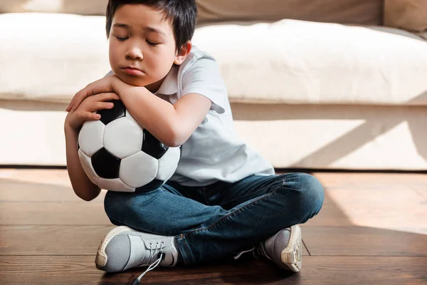Niño molesto con balón de rugby sentado en el suelo en casa en cuarentena. - foto de stock