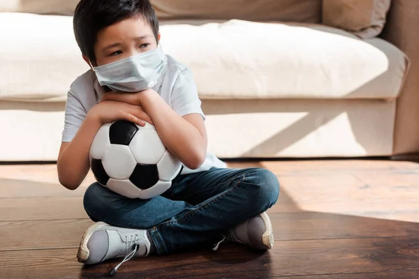 Triste enfant asiatique avec un ballon de football assis par terre à la maison en auto-isolement — Photo de stock