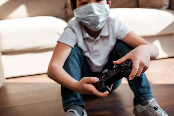 KYIV, UKRAINE - APRIL 22, 2020: asian boy in medical mask playing video game with joystick on quarantine — Stock Photo