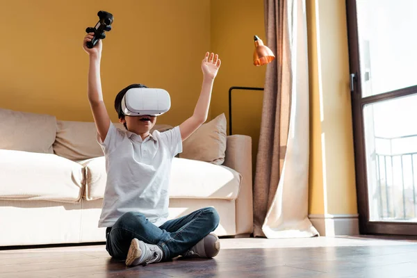 KYIV, UKRAINE - APRIL 22, 2020: jovencito excitado jugando video juego con joystick y la realidad virtual auriculares en auto aislamiento. - foto de stock