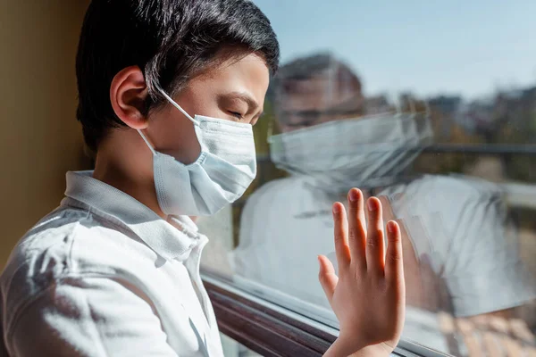 Upset asian child in medical mask looking through window on quarantine — Stock Photo