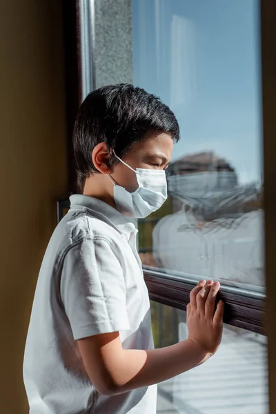 Мальчик в медицинской маске смотрит в окно на карантин — стоковое фото