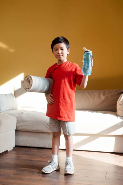 Niño asiático atlético con botella de deporte y colchón de fitness en casa durante el autoaislamiento. - foto de stock