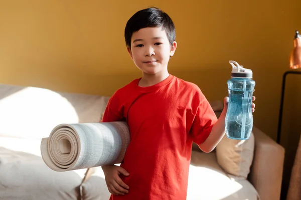 Спортивна азіатська дитина тримає спортивну пляшку і фітнес килимок вдома під час самоізоляції — стокове фото