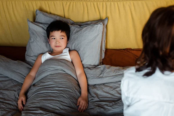 Chico asiático lindo acostado en cama con madre cerca de la cuarentena - foto de stock