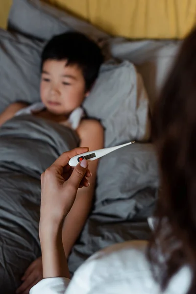 Ill asian boy lying in bed while mother holding thermometer on quarantine, selective focus — Stock Photo