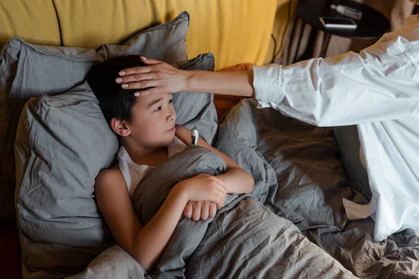 Chico asiático enfermo con termómetro acostado en la cama con madre cerca de la cuarentena. - foto de stock
