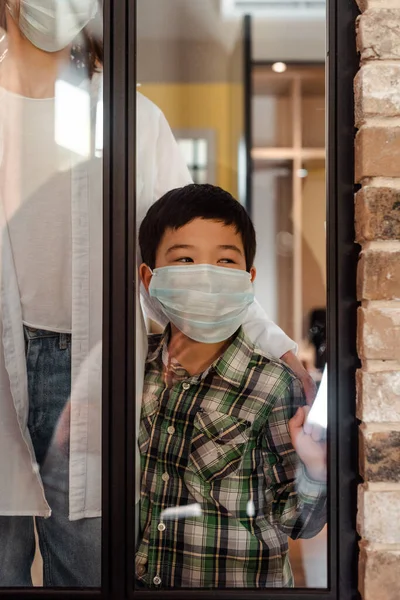 Asian boy in medical mask standing near window with mother on quarantine — Stock Photo