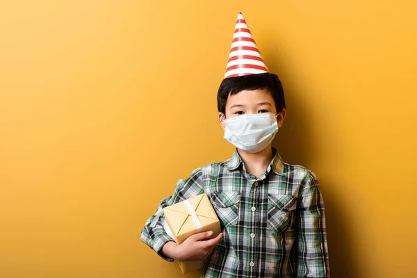 Asian boy in party cone and medical mask holding birthday gift on yellow during self isolation — Stock Photo