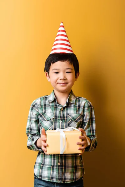 Chico asiático lindo en el cono de fiesta sosteniendo el regalo de cumpleaños en amarillo - foto de stock