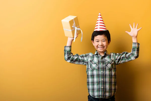 Happy asian boy in birthday party cone holding present on yellow — Stock Photo