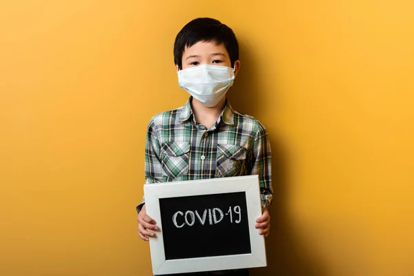 Asiatischer Junge in medizinischer Maske mit Covid-19-Schild auf gelb während der Quarantäne — Stockfoto