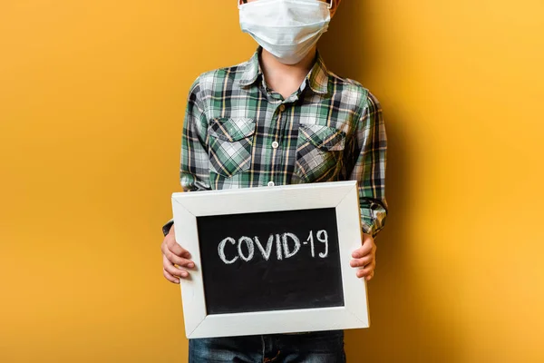 Ausgeschnittene Ansicht des Jungen in medizinischer Maske Haltebrett mit covid-19 Zeichen auf gelb während der Selbstisolation — Stockfoto