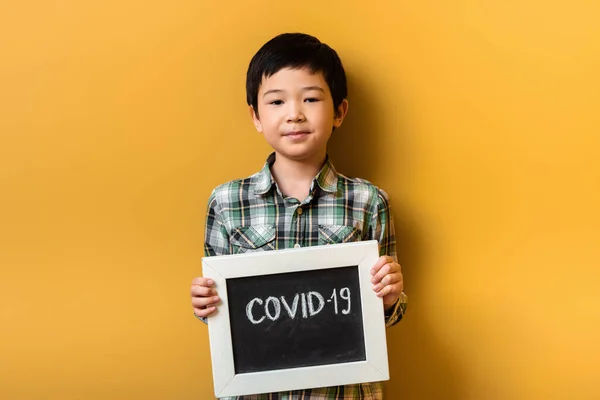 Lächelnder asiatischer Junge hält während der Quarantäne Schild mit Covid-19-Zeichen auf gelb — Stockfoto