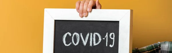 Cropped view of child holding board with covid-19 sign on yellow during self isolation, horizontal crop — Stock Photo