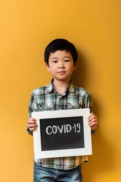 Asian boy holding board with covid-19 sign on yellow during quarantine — Stock Photo