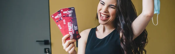 Panoramic shot of excited girl holding medical mask and night party tickets, end of quarantine concept — Stock Photo