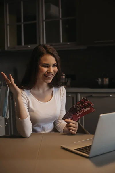 Mujer feliz celebración de boletos de fiesta de noche y máscara médica cerca de la computadora portátil, fin del concepto de cuarentena - foto de stock