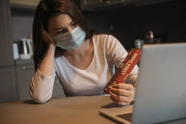 Foco seletivo de freelancer triste em máscara médica segurando bilhetes de festa noite perto de laptop em casa — Fotografia de Stock