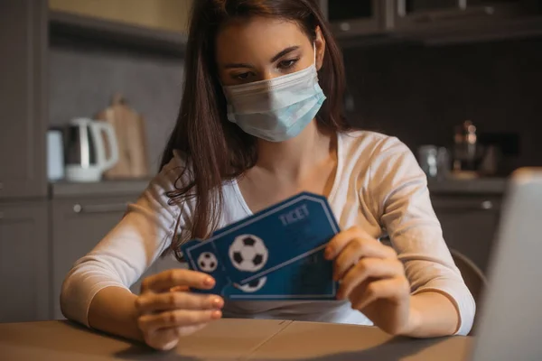Selective focus of sad woman in medical mask holding sport match tickets at home — Stock Photo