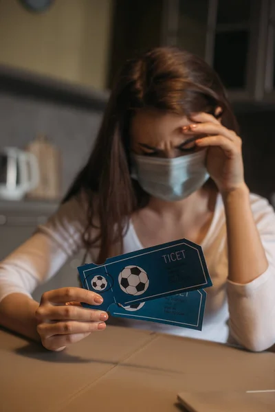 Mulher frustrada em máscara médica segurando bilhetes de jogo esporte em casa — Fotografia de Stock