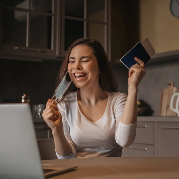 Fuoco selettivo di libero professionista felice in maschera medica che tiene il passaporto con biglietto aereo vicino a computer portatile, fine di concetto di quarantena — Foto stock