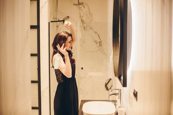 Mujer feliz en vestido negro hablando en el teléfono inteligente y la celebración de máscara médica, fin del concepto de cuarentena - foto de stock