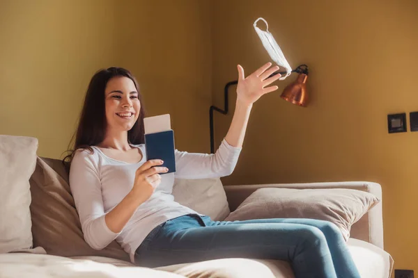 Alegre mujer lanzando en el aire máscara médica y la celebración de pasaporte con billete de avión mientras está sentado en el sofá, fin del concepto de cuarentena - foto de stock