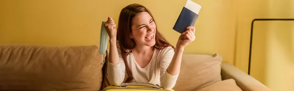 Orientação panorâmica da jovem feliz com máscara médica segurando passaporte e bilhete de ar, fim do conceito de quarentena — Fotografia de Stock