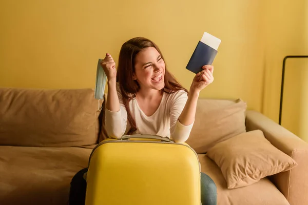 Excited young woman with medical mask holding passport and air ticket near baggage, end of quarantine concept — Stock Photo