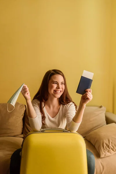Happy young woman with medical mask holding passport and air ticket near luggage, end of quarantine concept — Stock Photo
