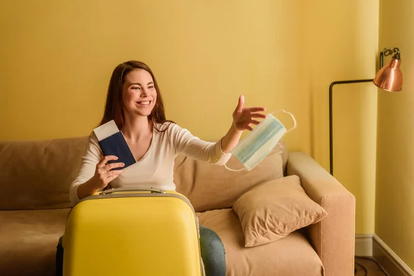 Feliz joven mujer lanzando en el aire máscara médica y la celebración de pasaporte con billete de avión cerca de equipaje, fin del concepto de cuarentena — Stock Photo