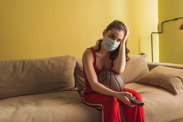 Спортсменка в медицинской маске держит пульт дистанционного управления возле баскетбола во время просмотра чемпионата в гостиной — стоковое фото