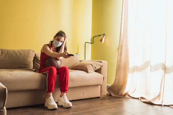 Sportive girl in medical mask holding remote controller near basketball while watching championship in living room — Stock Photo