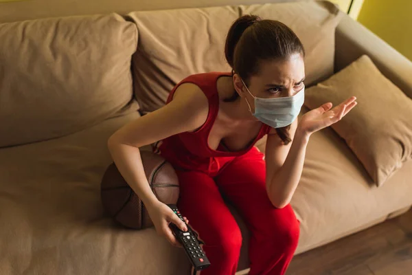 Ansicht der emotionalen Sportlerin in medizinischer Maske mit Fernbedienung in der Nähe von Basketball, während sie die Meisterschaft im Wohnzimmer verfolgt — Stockfoto