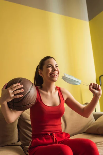 Desportista alegre tocando máscara médica e segurando basquete na sala de estar, fim do conceito de quarentena — Fotografia de Stock