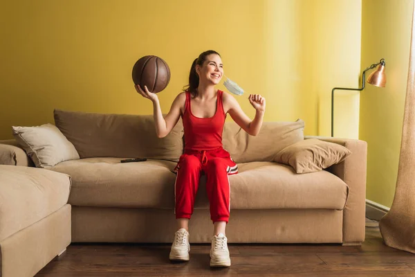 Glückliche Sportlerin berührt medizinische Maske und hält Basketball im Wohnzimmer, Ende der Quarantäne — Stockfoto