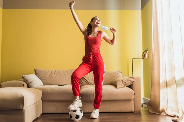 Glückliche Sportlerin mit Hand über dem Kopf, die medizinische Maske berührt und neben Fußball im Wohnzimmer steht, Ende der Quarantäne — Stockfoto